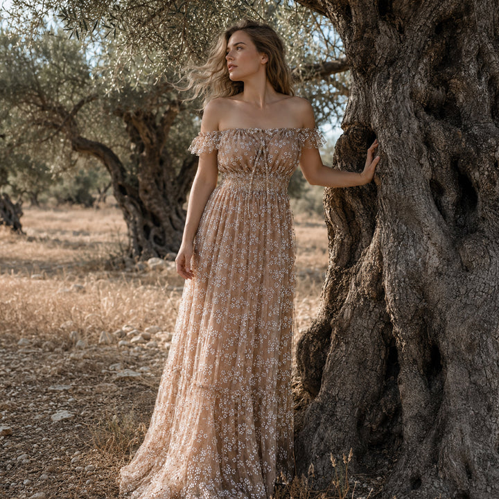 A woman stands beside a gnarled olive tree in a sun-dappled grove, wearing a sheer khaki off-the-shoulder tulle gown with delicate white floral embroidery and ruffled short sleeves that flows to the rocky ground.