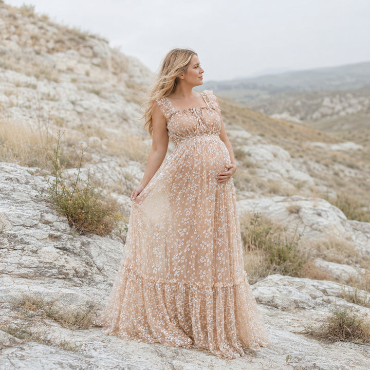 A pregnant woman stands on a rocky outcrop in a rugged, hilly landscape wearing a flowing beige floral embroidered tulle maternity gown with ruffled cap sleeves and a tiered skirt that drapes over the stones.