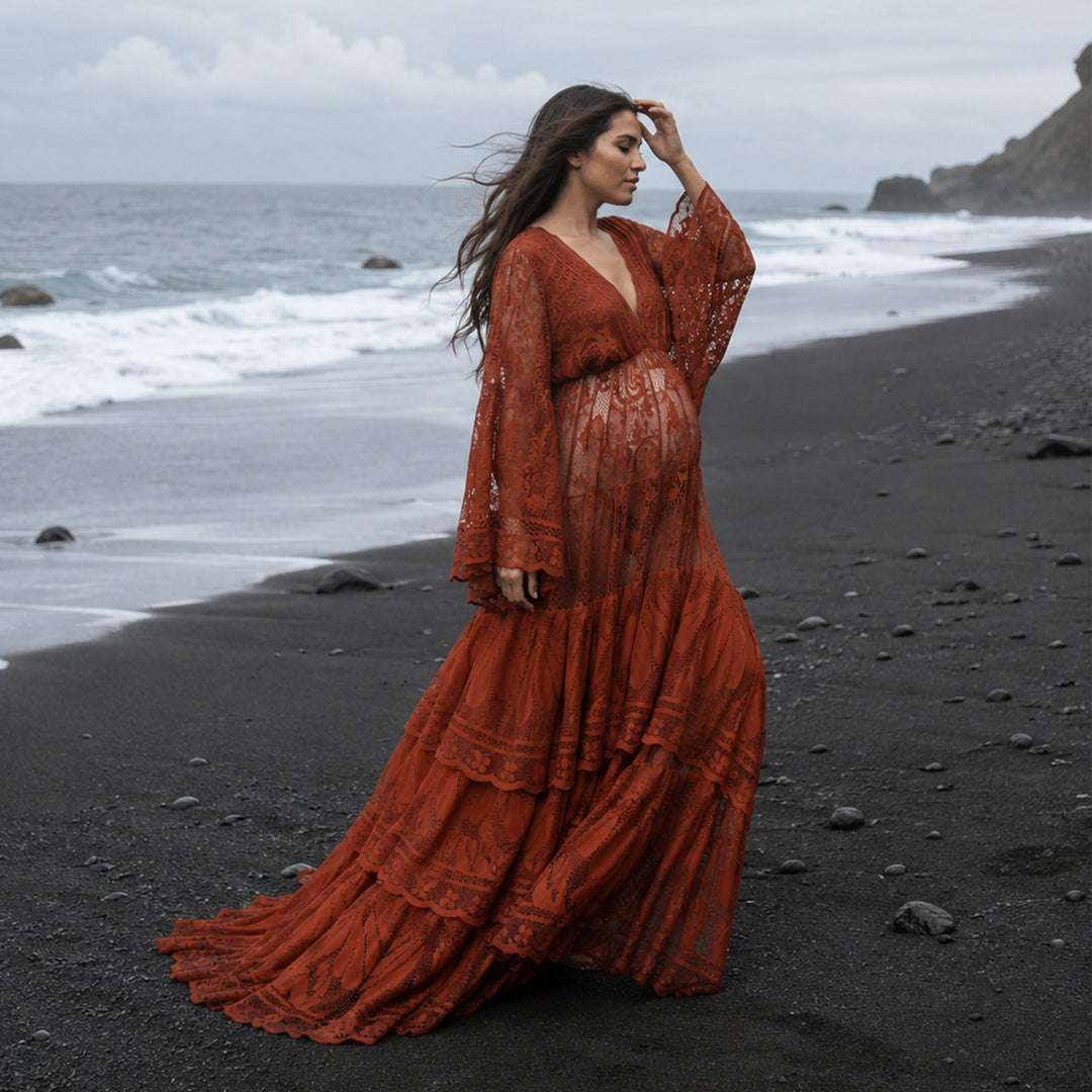 A pregnant woman stands in profile on a rocky black beach near the water's edge, wearing a long terracotta lace dress with long sleeves. 