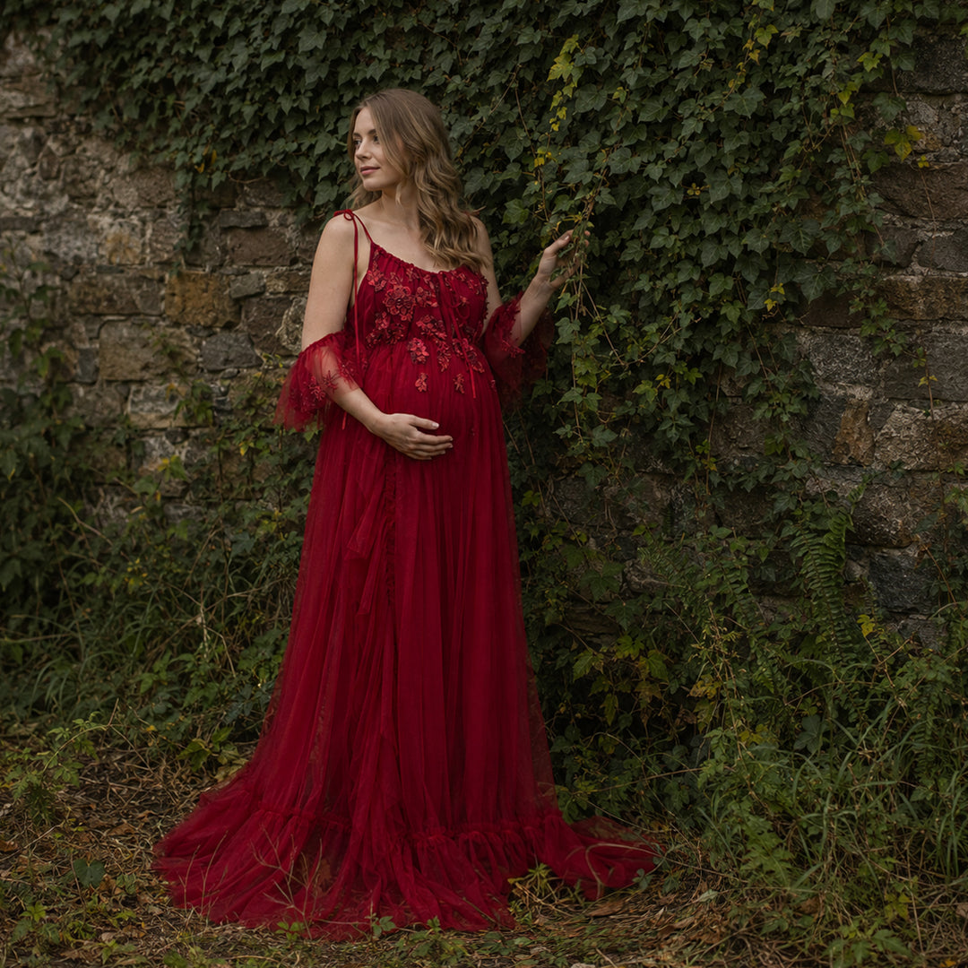 A pregnant woman wears red dress with 3D floral embroidery and ruffled sleeves, it's train trailing on the ground; she touches ivy with her left hand, cradles her belly with her right, standing.
