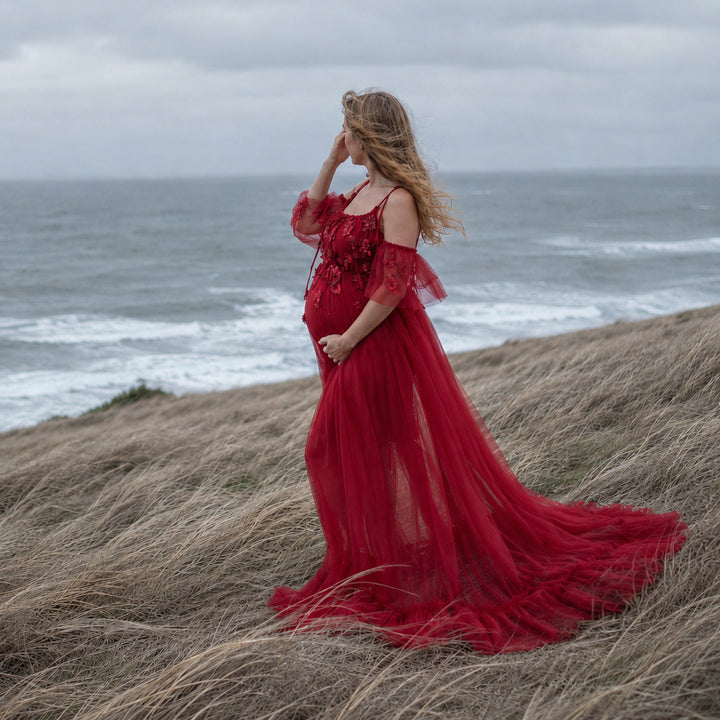 A pregnant woman in a red spaghetti-strap gown with a trailing skirt stands on a grassy slope; she touches her hair with her right hand, cradles her belly with her left, with the sea and waves in the background.