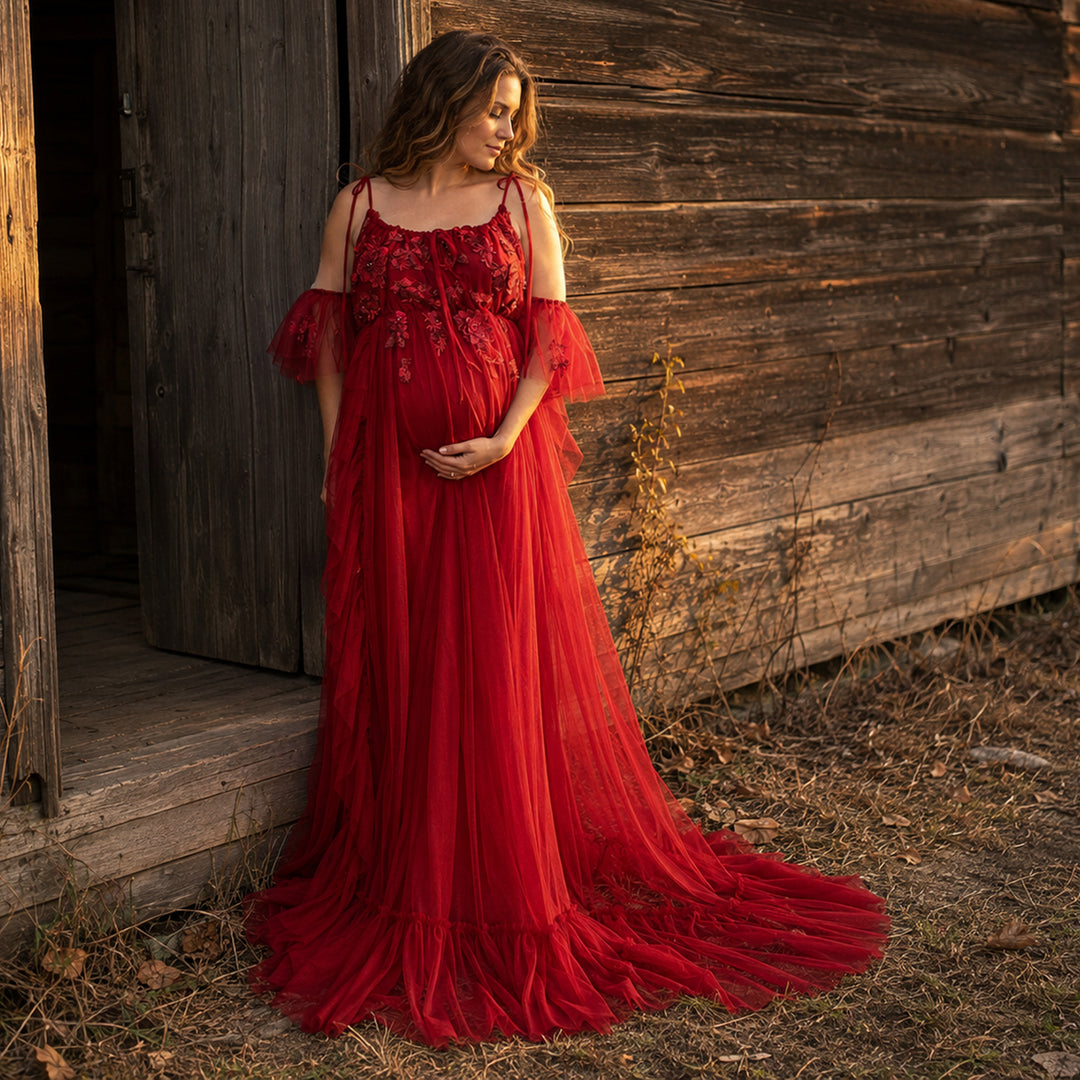  pregnant woman in a red spaghetti-strap gown with floral embroidery, ruffled sleeves and a trailing skirt stands before a wooden cabin; she cradles her belly with both hands, looking down.
