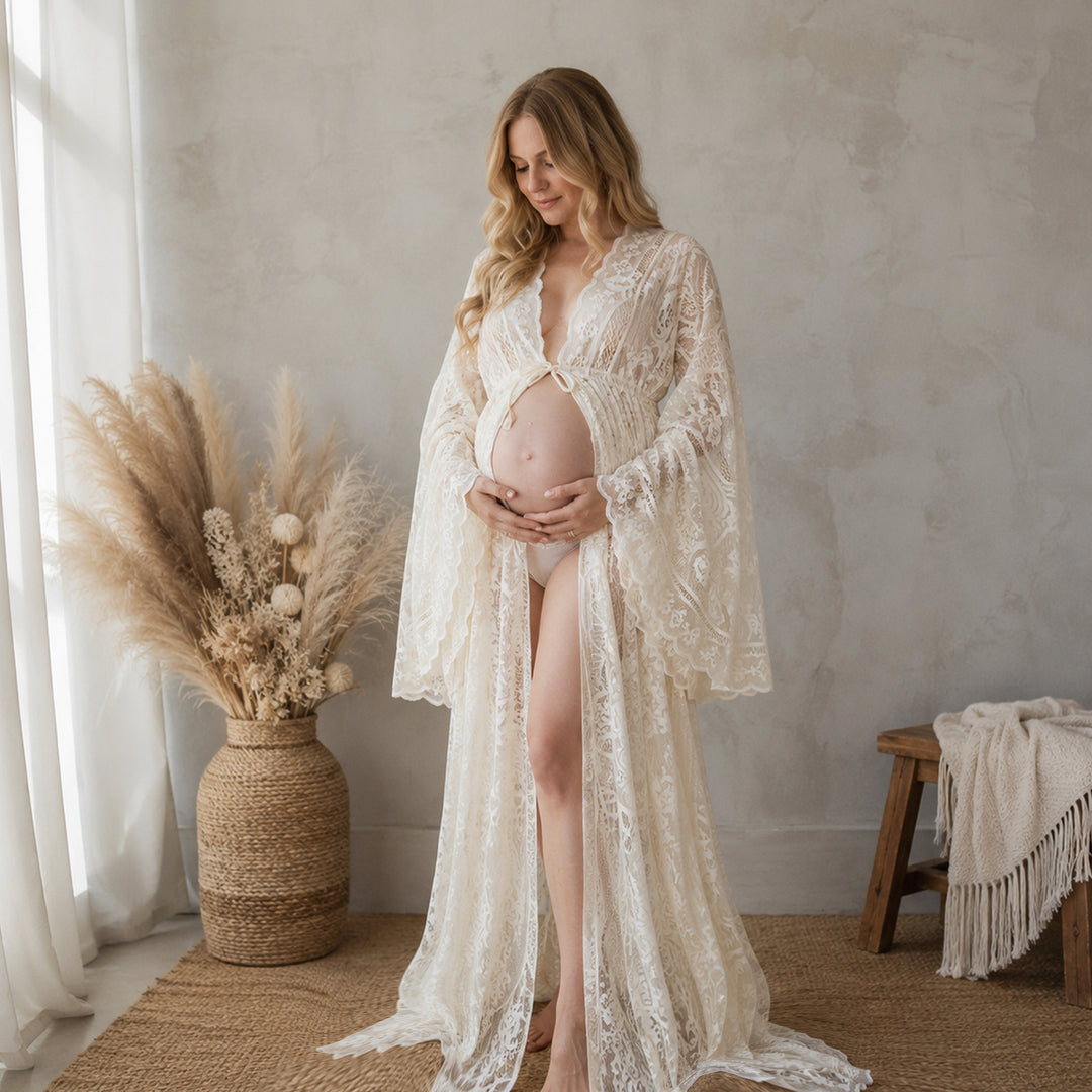 A pregnant woman stands indoors on a woven rug wearing a beige lace maternity robe with long bell sleeves and an open front.